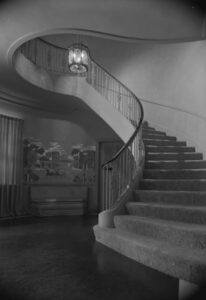 Photograph of an interior view of the Chi Omega Sorority House. There is a carpeted spiral staircase, and a mural on the wall in the background with a hanging light in the center of the foyer.