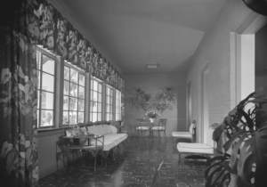 Photograph of an interior view of the Chi Omega Sorority House. There are exterior windows with floral curtains, and couches and chairs around the hallway.