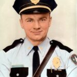 man in blue uniform with black trim and pocket tops wearing a black policeman's hat.