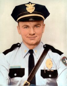 man in blue uniform with black trim and pocket tops wearing a black policeman's hat.