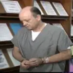 prisoner in handcuffs walking by shelf with stacks of paper